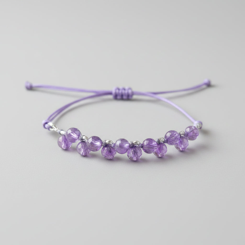 A purple cord bracelet with faceted purple beads and small silver spacer rings, resting on a textured gray fabric surface.