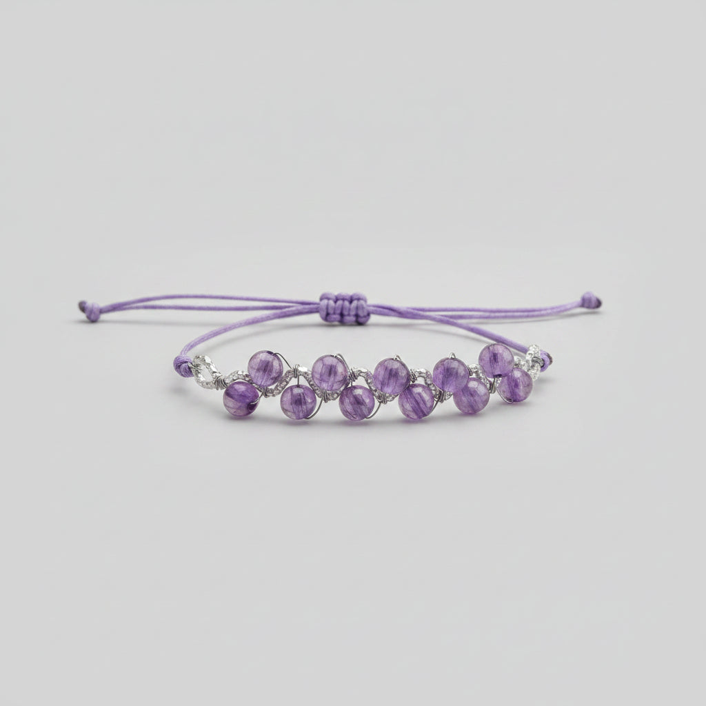 A purple beaded bracelet with faceted beads and silver-toned metal accents, resting on a textured gray fabric surface with a blurred indoor background.