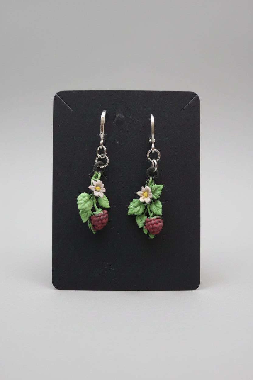 A pair of handmade resin earrings shaped like green leaves and red raspberries with small white flowers, hanging from silver fishhook wires on a black display card, set against a gray carpeted background.