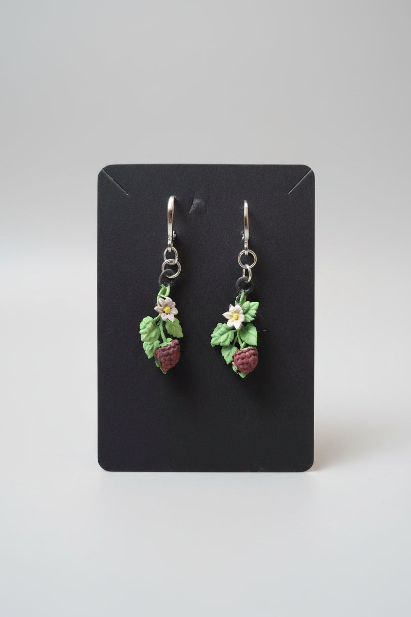 A pair of handmade dangle earrings featuring small green leaves, white flowers, and red raspberries hanging from silver fishhook wires, displayed on a black backing card over a gray textured surface.