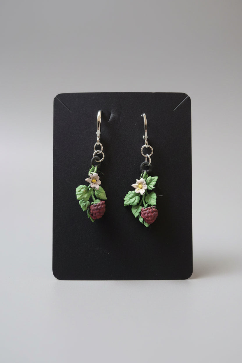 A pair of handmade resin dangle earrings featuring green leaves, small white flowers, and red raspberries, displayed on a black card against a gray textured background.