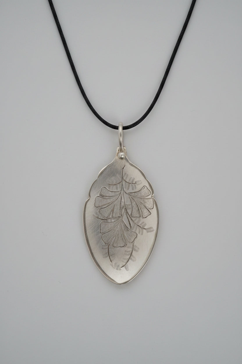 A silver-toned pendant necklace with an engraved leaf design, hanging from a black cord against a textured gray fabric background.