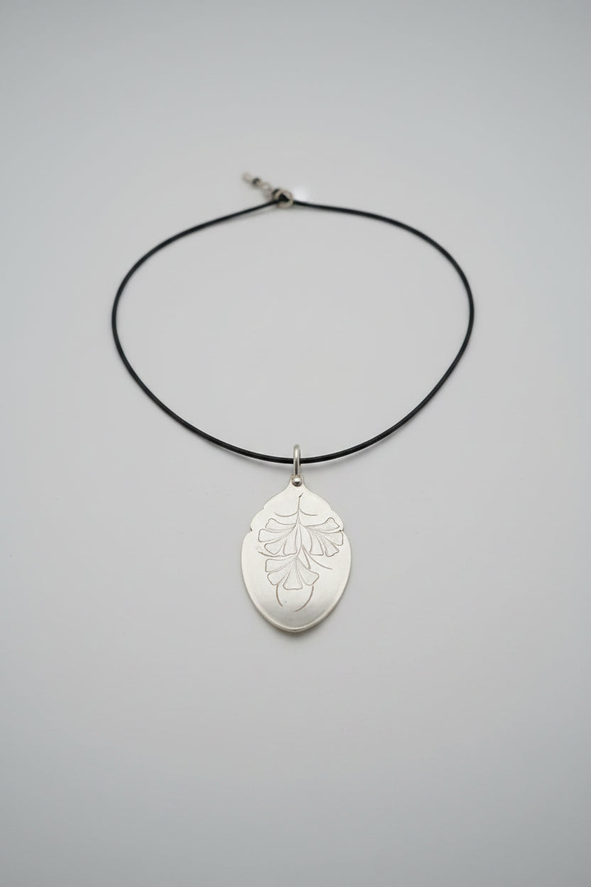 A silver pendant necklace featuring an engraved leaf design on a black cord, displayed on a textured gray fabric surface.