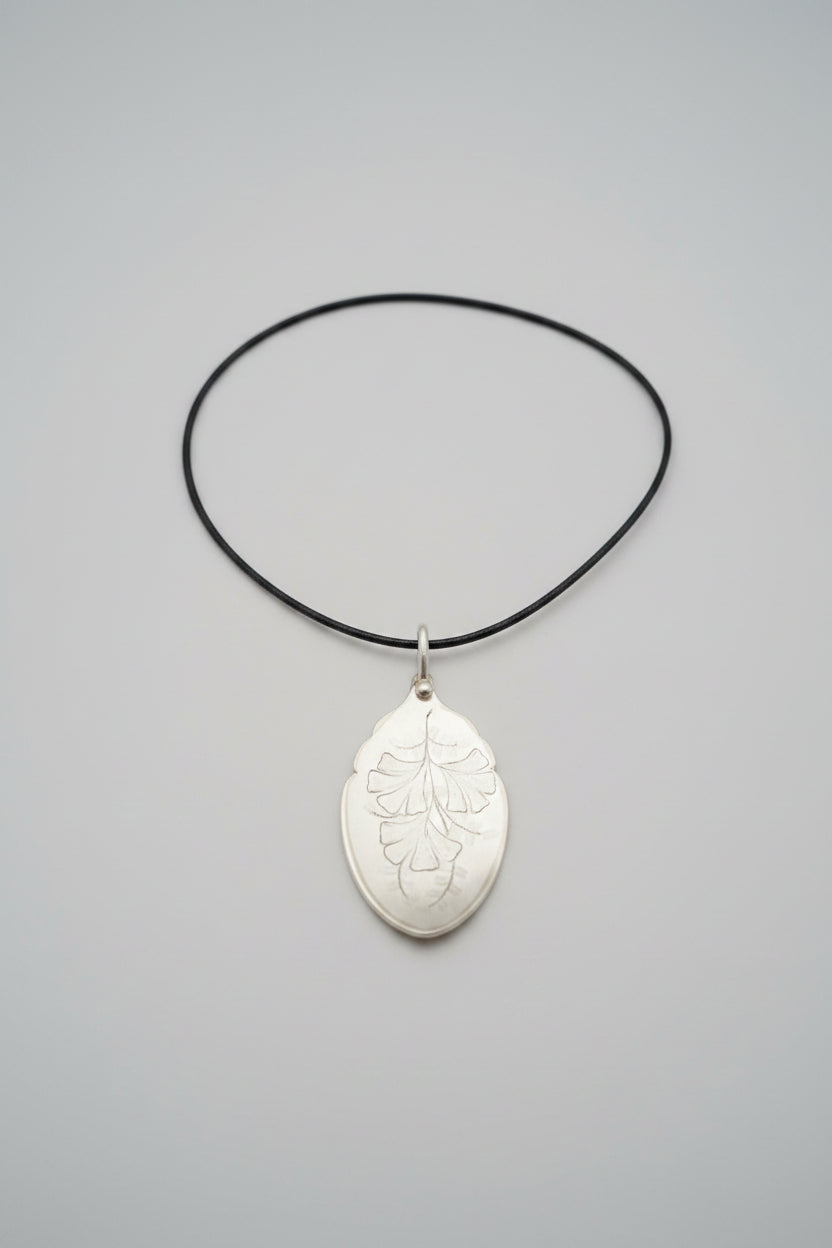 A silver-toned pendant necklace featuring an engraved leaf design on a black cord, displayed on a textured gray fabric background.