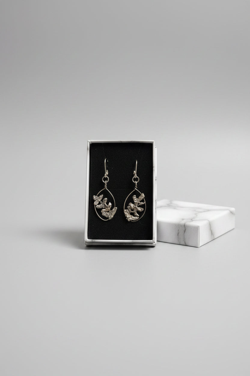 A pair of silver drop earrings with leaf-like filigree details, presented in a black velvet box held by a hand in a home setting.