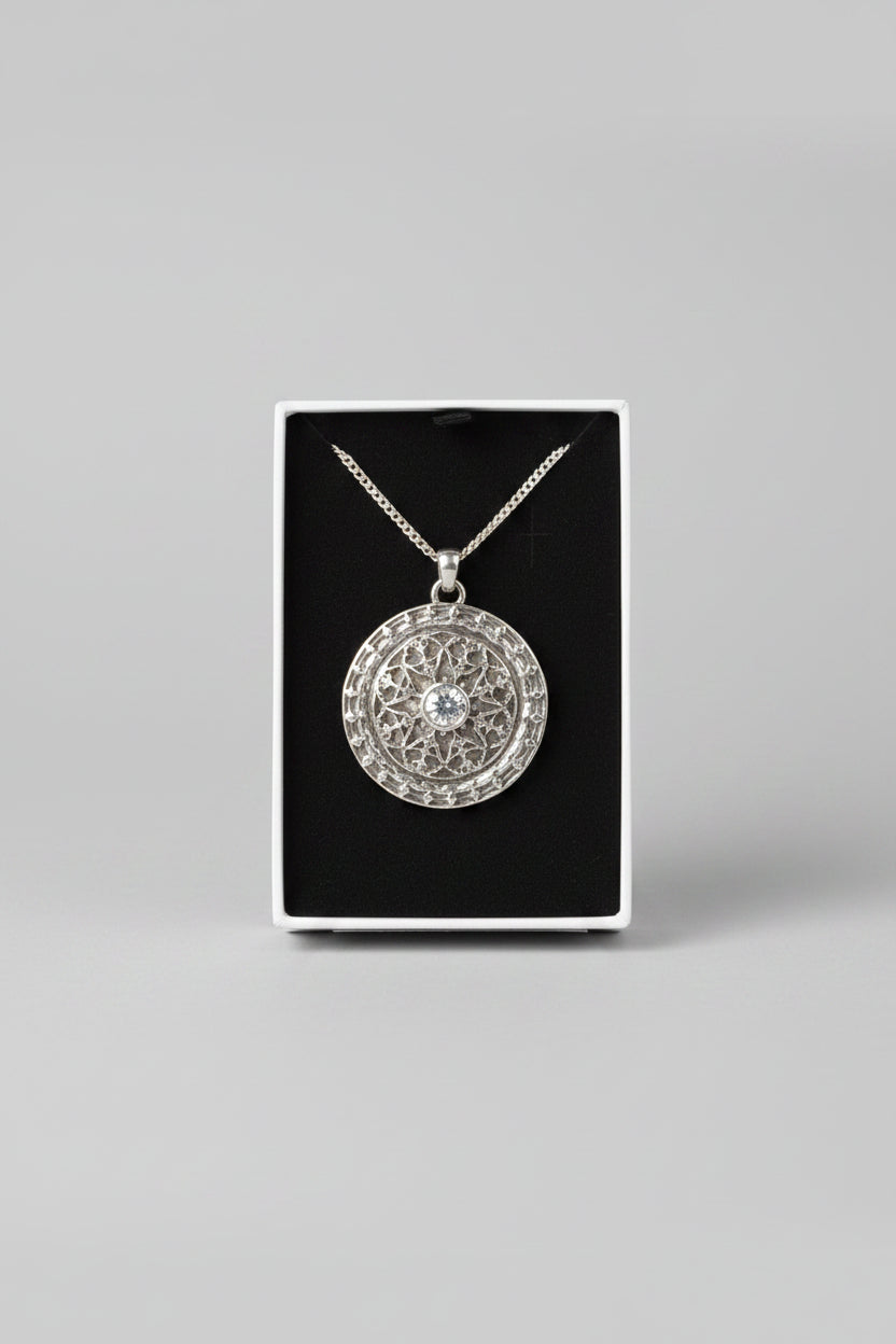 A hand holding a black jewelry box containing a round textured pendant necklace on a chain, with a laptop blurred in the background.