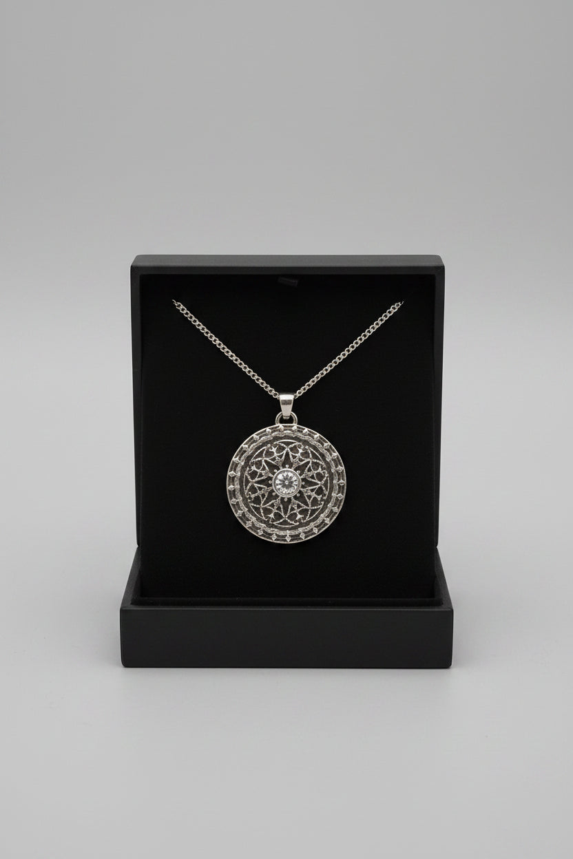 A round silver pendant necklace with an intricate filigree pattern and a central clear stone, displayed on a black velvet insert inside an open box held by a hand, with a laptop in the background.