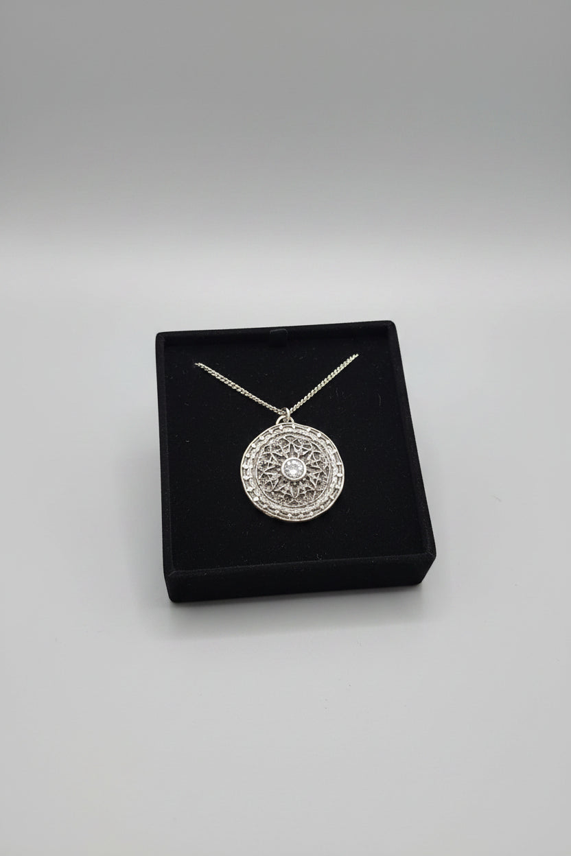 A hand holding a silver-toned circular pendant necklace on a black display card inside a small box, with a blurred laptop in the background.