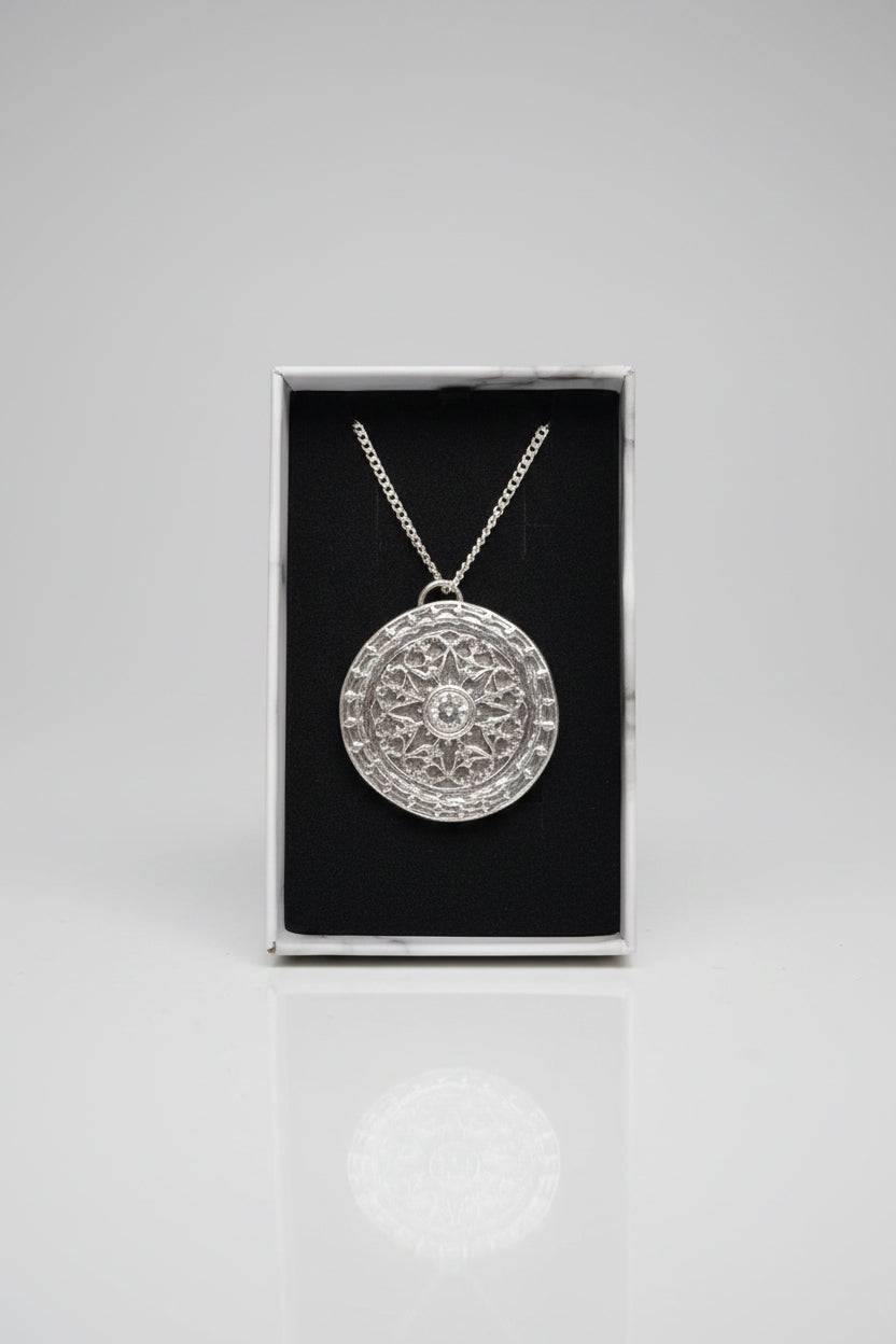 A silver pendant necklace with an intricate circular cutout design hangs from a silver chain inside an open jewelry box with a black insert. The box is placed on top of a black and white display tray.