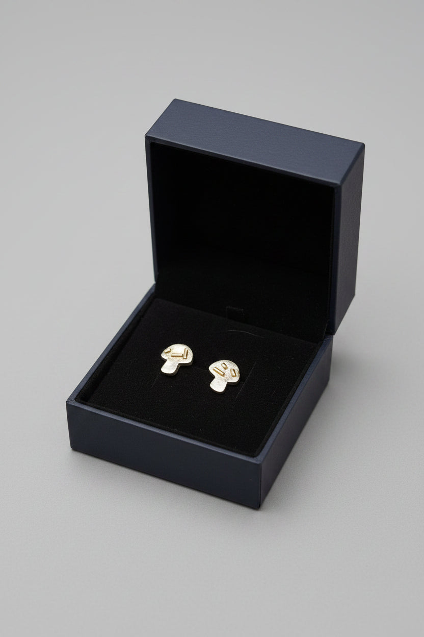 A pair of small mushroom-shaped stud earrings in a black presentation box on a textured gray carpet background.