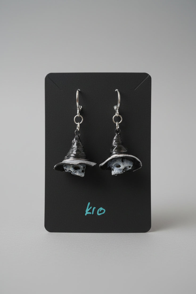 A pair of dangling witch hat earrings with skull details, displayed on a black card held by a hand. The earrings feature silver hooks and dark, weathered black and gray hats with white skull faces.