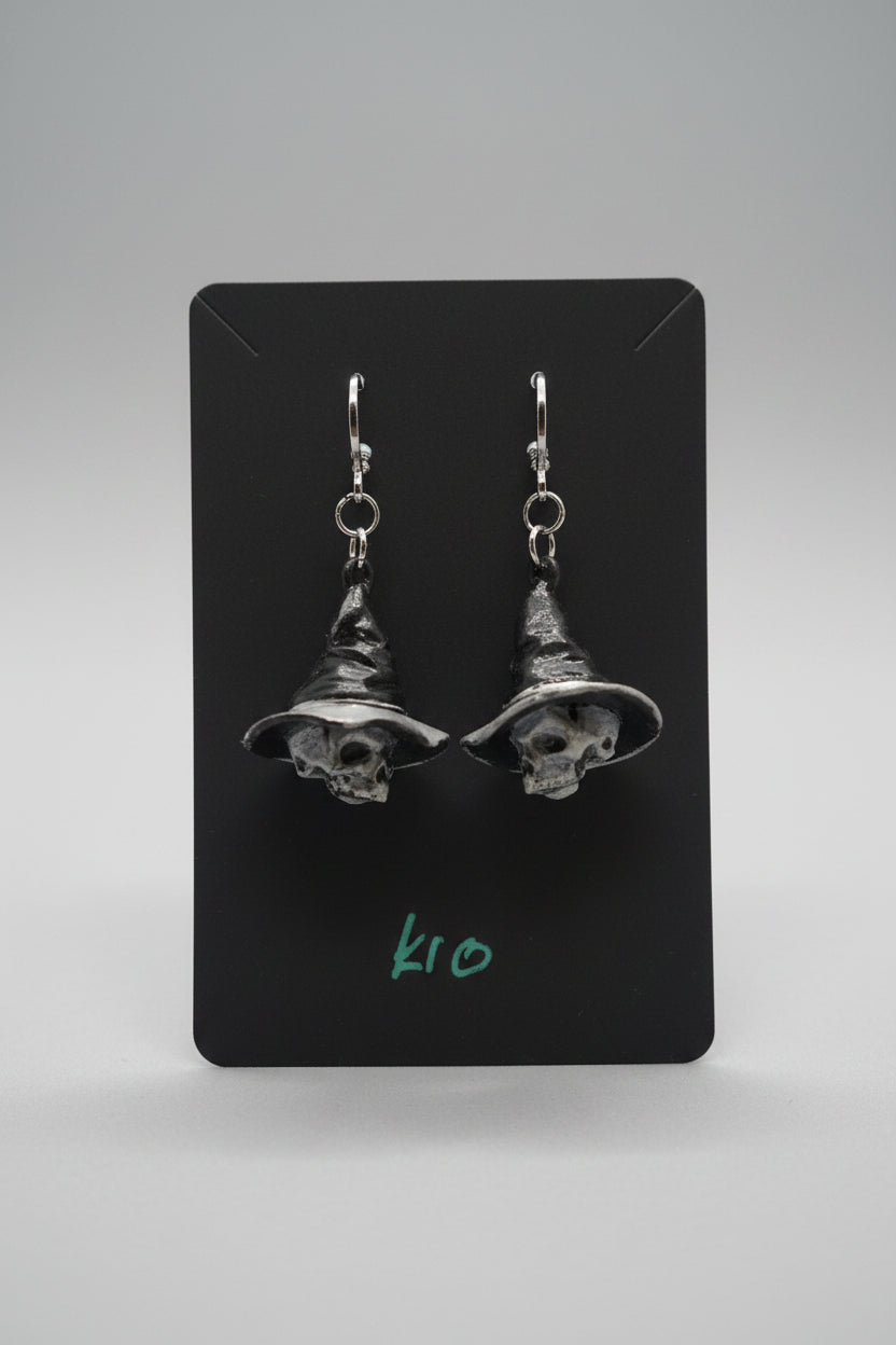 A pair of dangling witch hat earrings displayed on a black card, held by a hand over a gray textured fabric surface in an indoor setting.