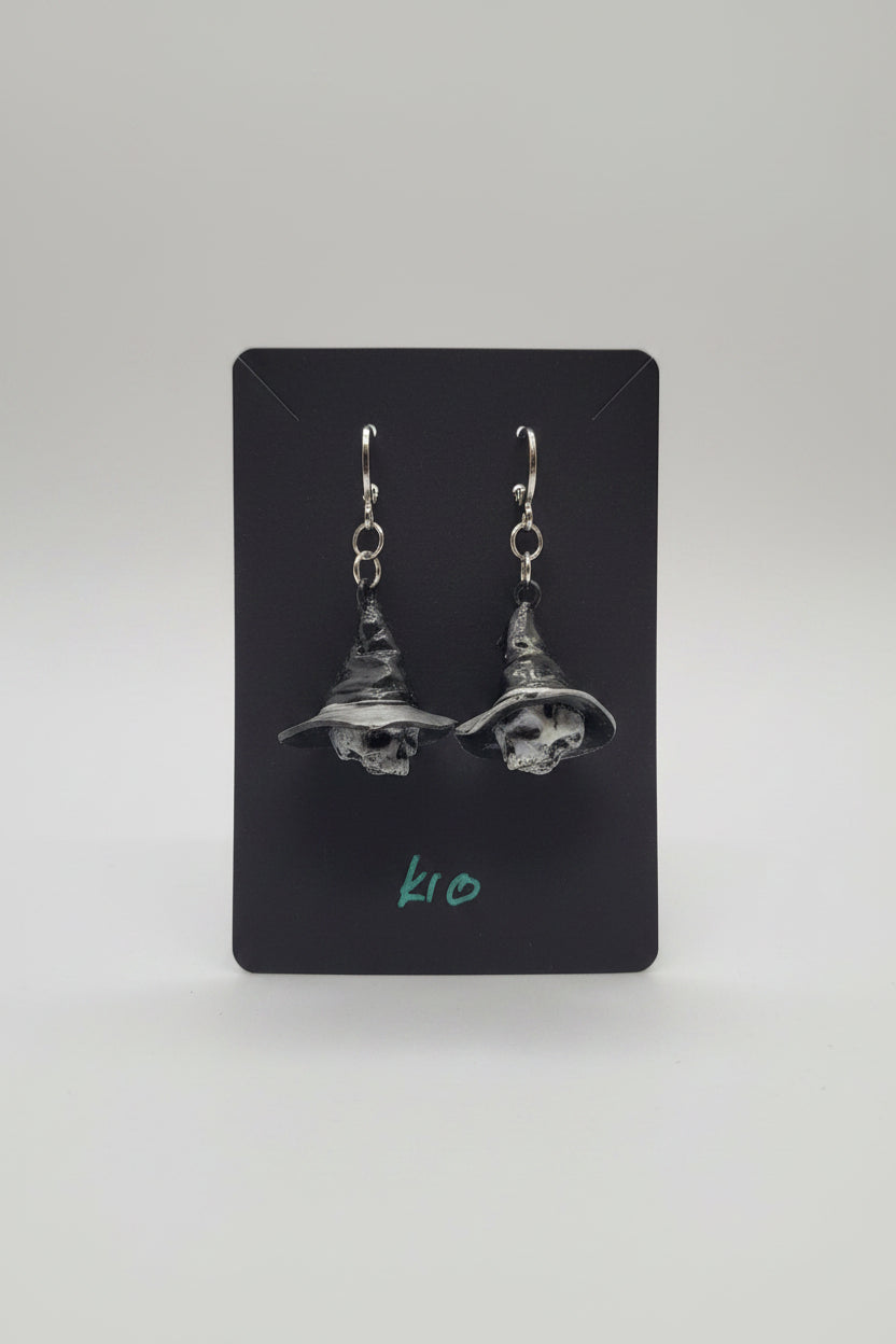 A pair of dangling witch hat earrings with skull details on a black display card, held by a hand in a home setting.