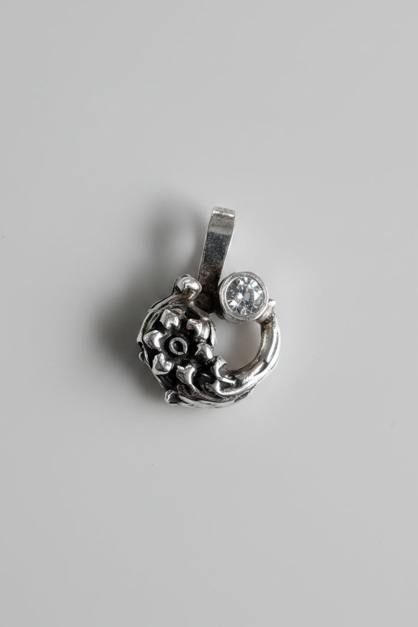 A small silver pendant with an ornate floral or scroll design and a clear round stone, shown on a textured beige fabric background.