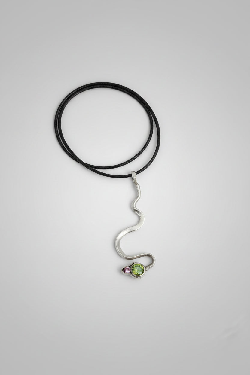 A silver snake pendant necklace with a small green gemstone at its head, presented inside a black box with a white marble-patterned border.