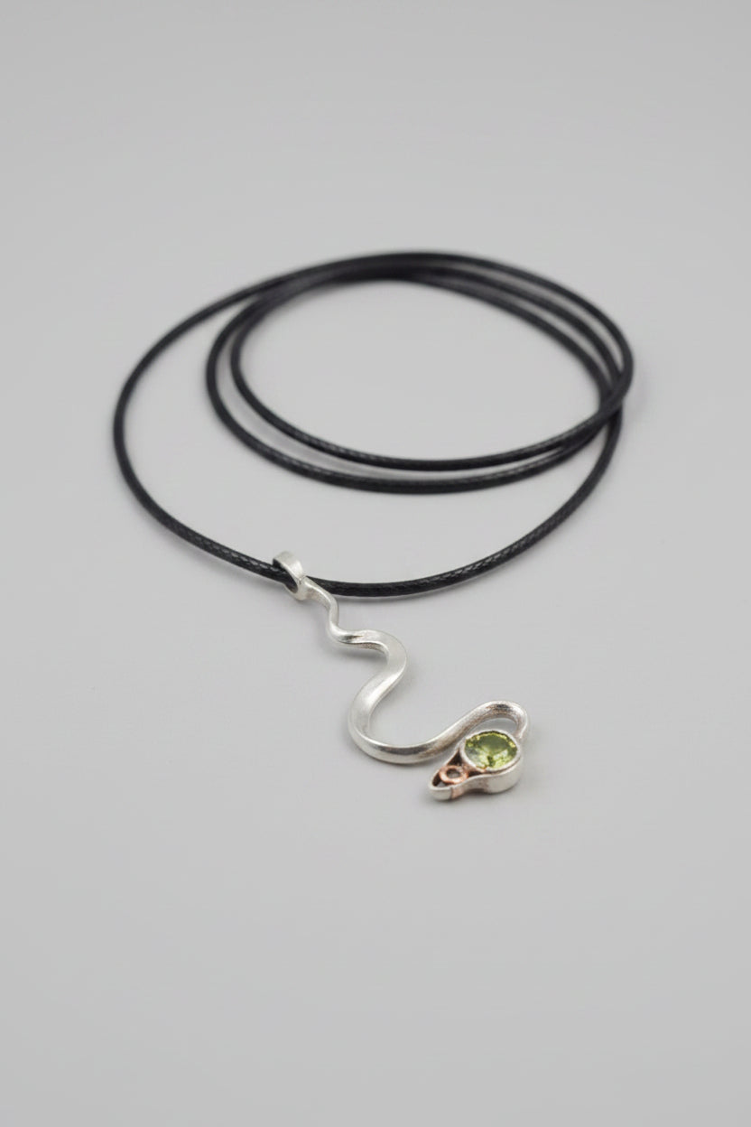 A silver snake pendant necklace with a green gemstone at its head, presented inside a gray marble-patterned box held by a hand.