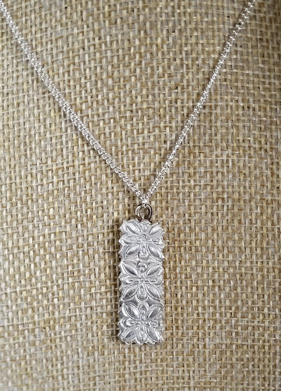 A silver rectangular pendant necklace with a floral pattern, displayed on a textured burlap background.