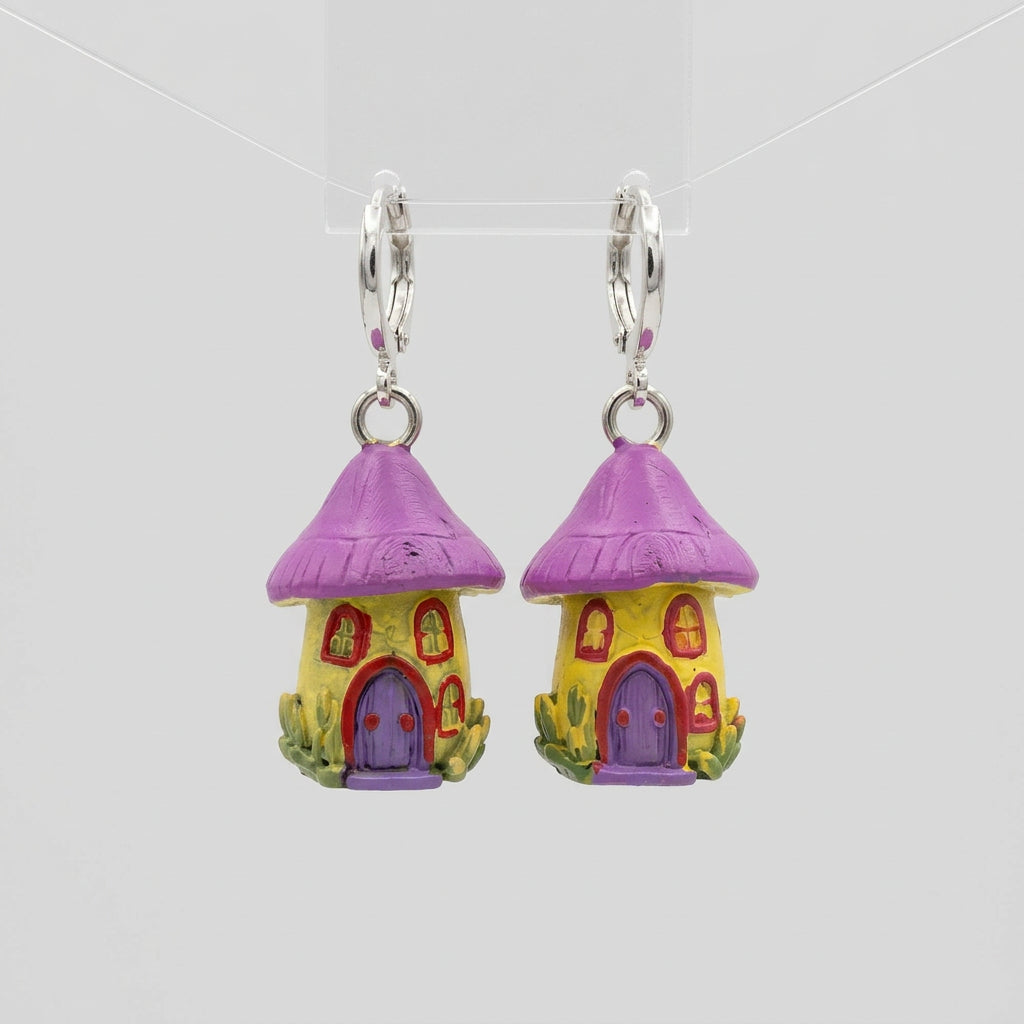 A pair of small dangling earrings shaped like tiny colorful houses, photographed on a textured gray fabric surface. Each earring has a silver-tone loop at the top and a small house charm with a pink-purple roof, yellow body, and purple door.