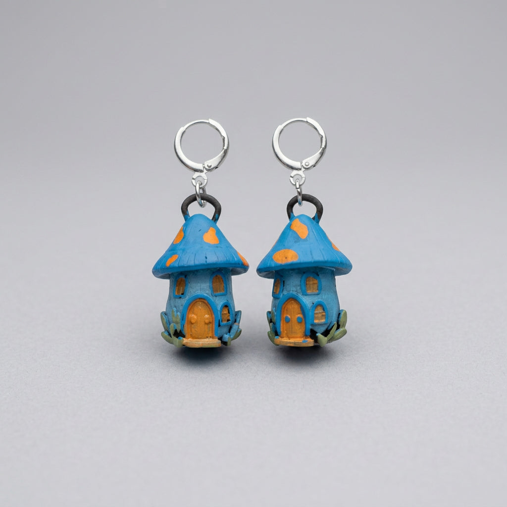 A pair of small fairy house charms with blue roofs, orange spots, and orange doors, attached to silver split rings, displayed on a gray textured fabric background.