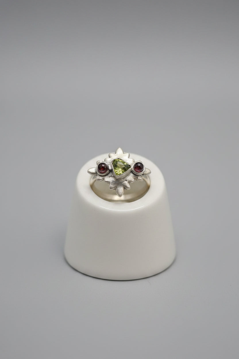 A silver ring with a floral cluster setting featuring a green center stone and red side stones, displayed on a black-lined wooden jewelry tray.