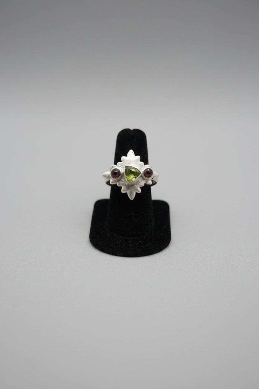 A silver ring with a green and red stone cluster setting, displayed on a black cushion inside a wooden jewelry organizer tray.