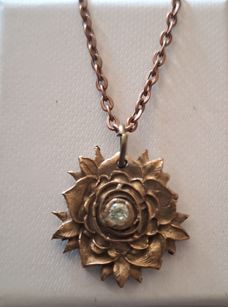 A rose-shaped pendant necklace in a warm bronze or copper-toned metal, featuring a small clear stone set in the center, displayed on a textured off-white background.