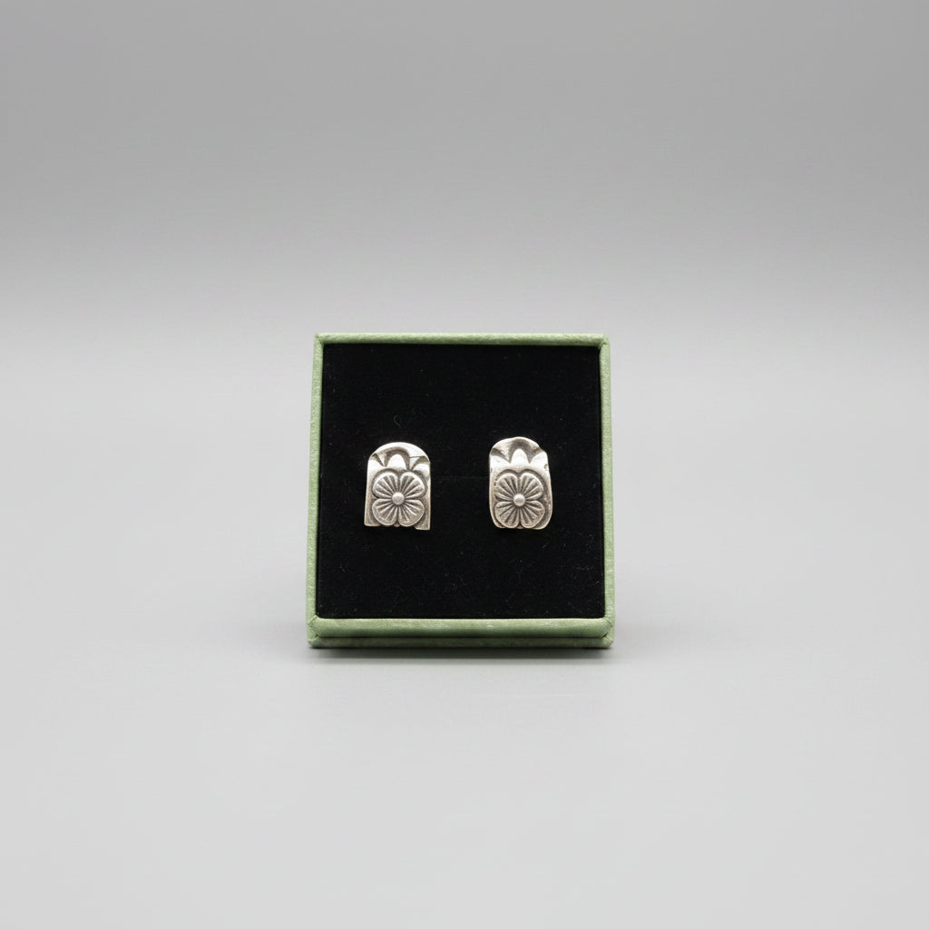 A pair of silver floral stud earrings displayed in a black presentation box with a green border, resting on a textured gray fabric surface.