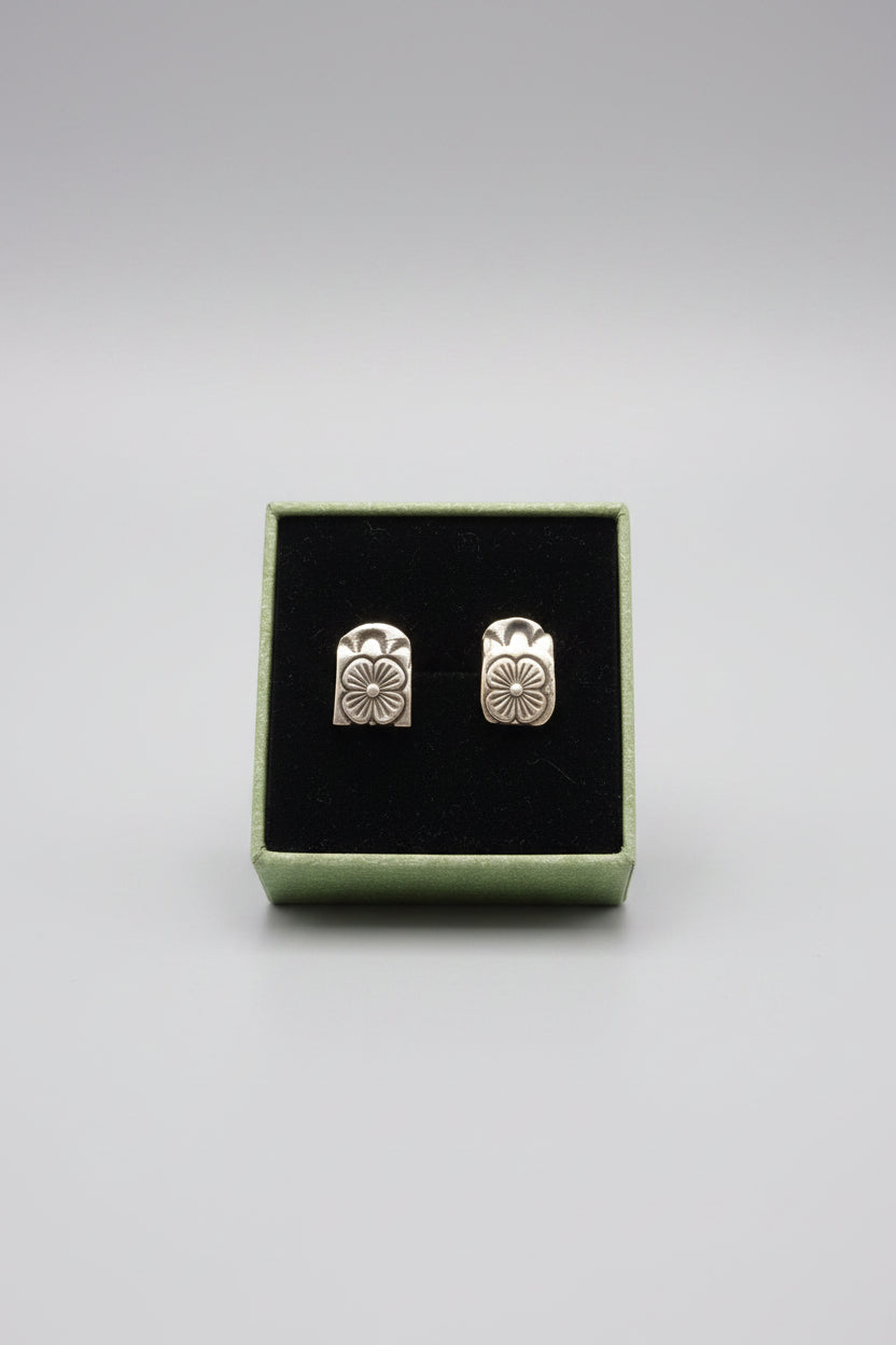 A pair of gold-toned flower-shaped stud earrings displayed in a black presentation box with a light green border, placed on a textured gray surface.