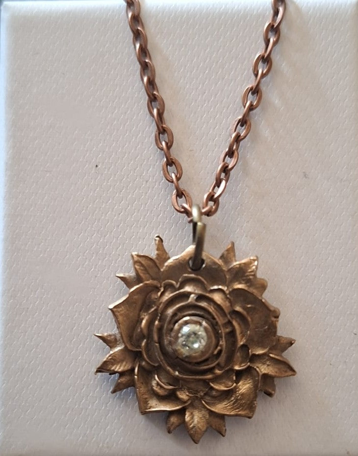 A rose-shaped pendant necklace in a warm bronze or copper-toned metal, featuring a small clear faceted gemstone in the center, displayed on a textured off-white background.