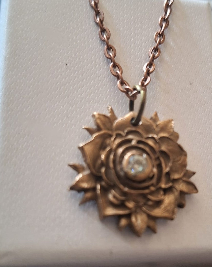 A rose gold flower pendant necklace with a central clear rhinestone, displayed on a textured white jewelry box.