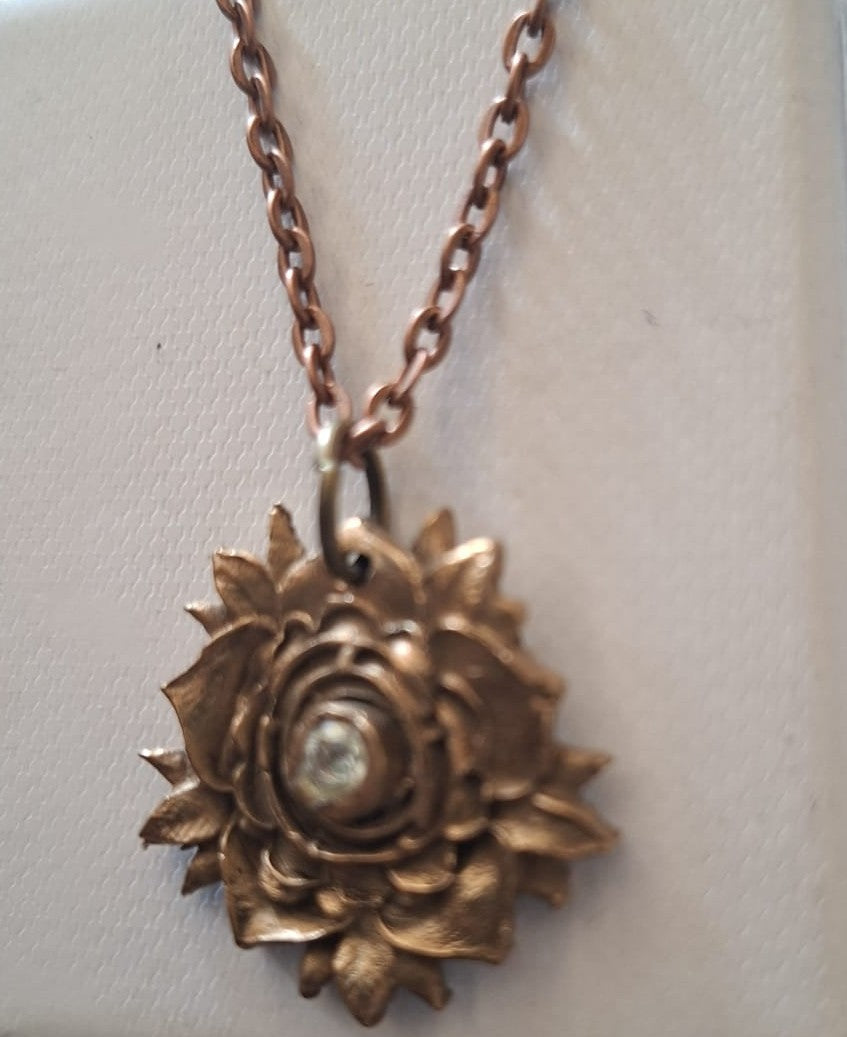 A close-up of a bronze-toned flower pendant necklace on a matching chain, displayed against a light beige textured background.
