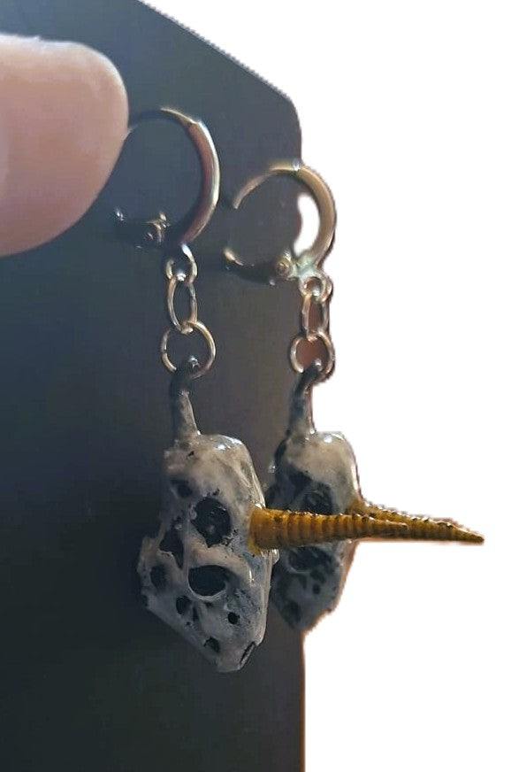 A pair of gothic dangle earrings featuring skull charms with embedded gold spiral accents, held by a finger against a black display card.