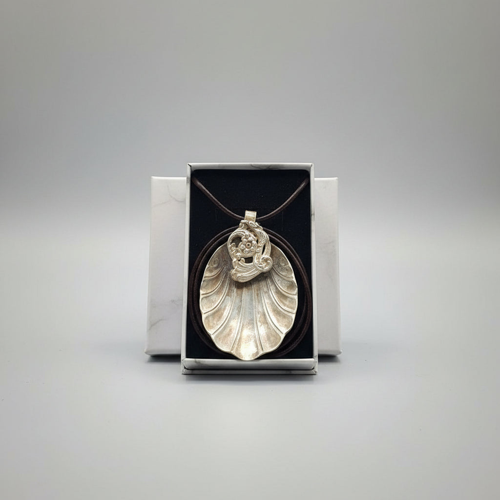 A silver shell-shaped pendant necklace with a detailed top knot, presented in an open silver box on a textured gray fabric surface.