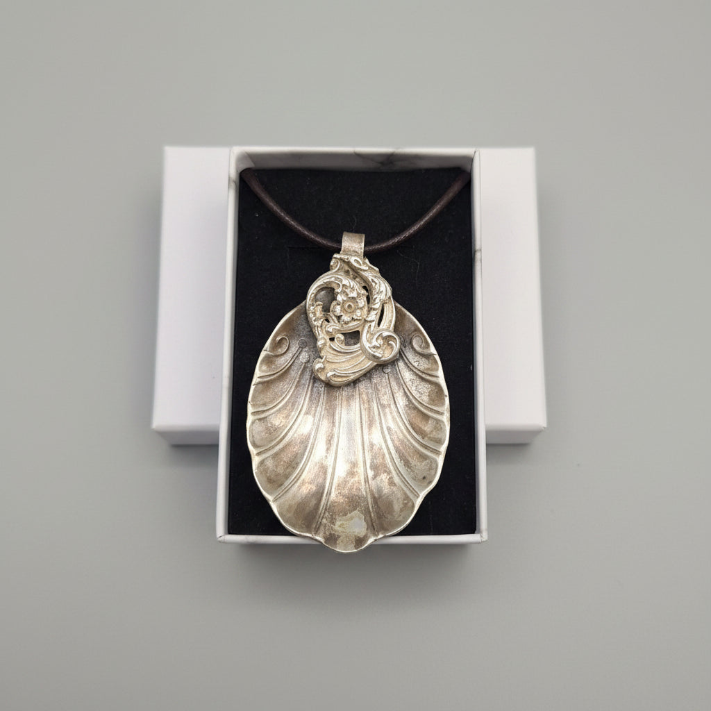 A silver-toned shell-shaped pendant necklace with an ornate figure design, displayed inside its open presentation box on a textured gray fabric surface.