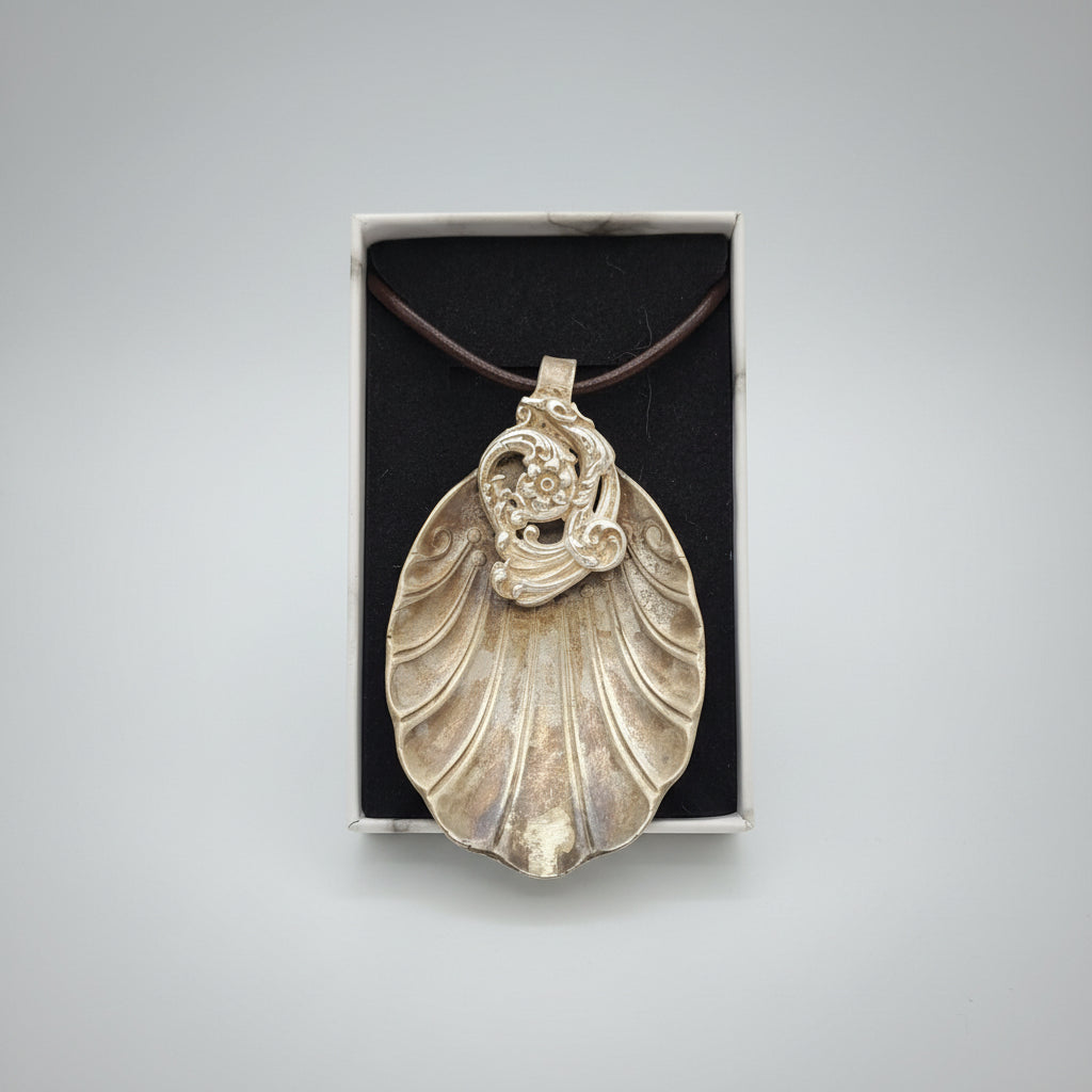 A silver-toned shell-shaped pendant necklace with a raised floral detail at the top, displayed inside an open rectangular gift box with a black insert. The necklace is attached to a dark cord, and the box is placed on a textured gray surface.