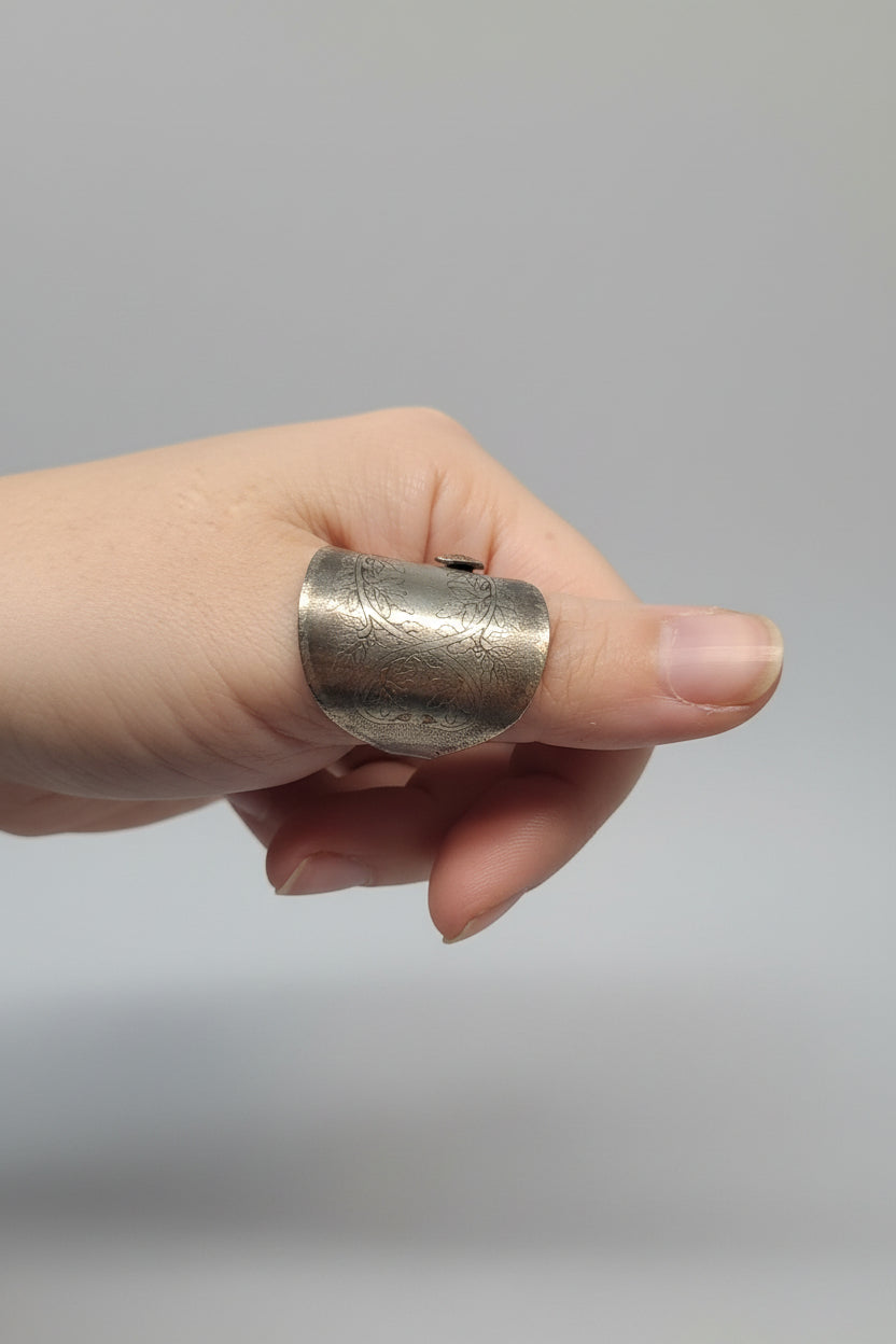 A handmade silver ring with a textured, hammered finish is shown worn on a person's finger, with a blurry indoor background.