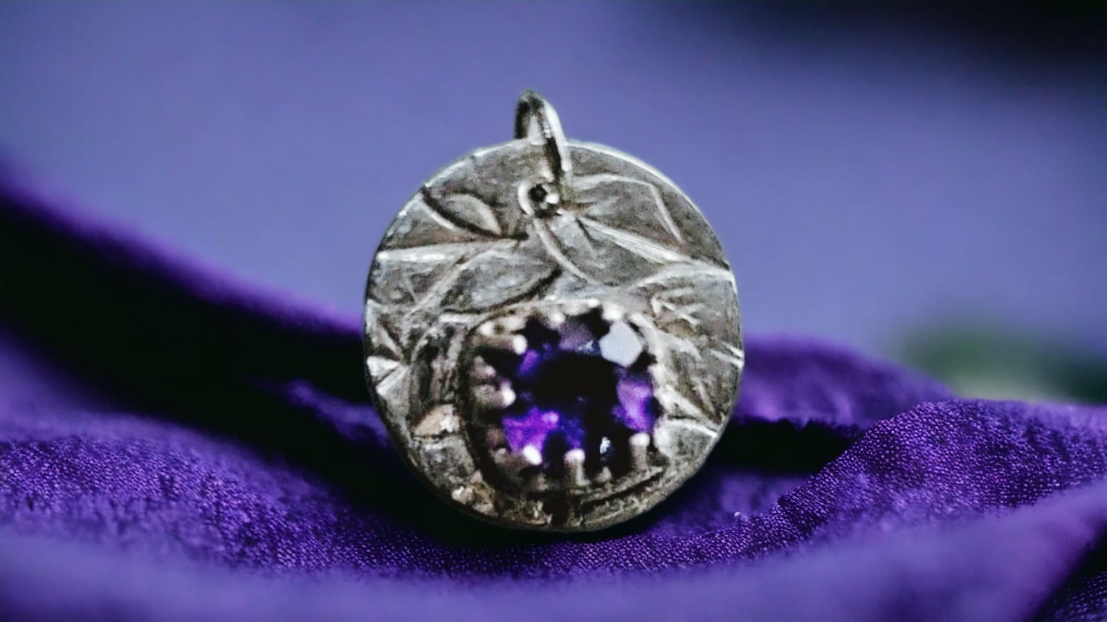 A silver-toned round pendant with a textured leaf-like border and a central purple gemstone, resting on draped purple fabric.