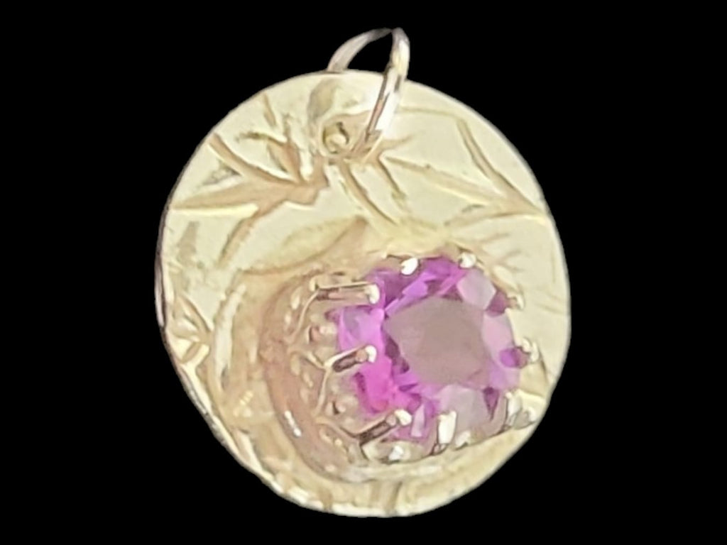 A round gold pendant featuring a textured, engraved design with a central bezel-set pink gemstone, shown against a solid black background.