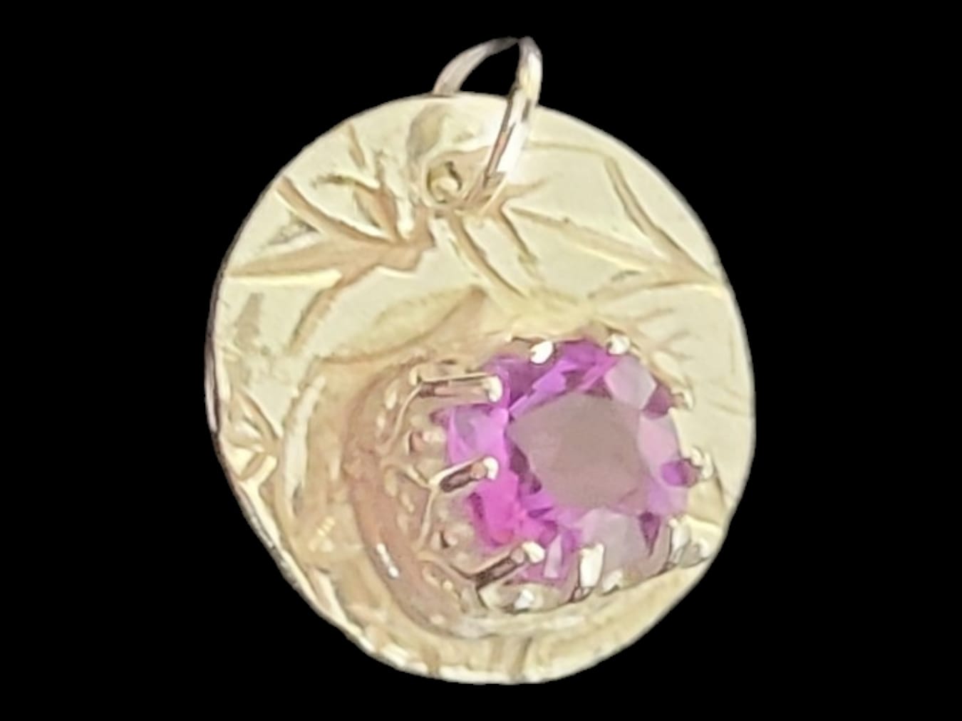 A round gold pendant featuring a textured, engraved design with a central bezel-set pink gemstone, shown against a solid black background.