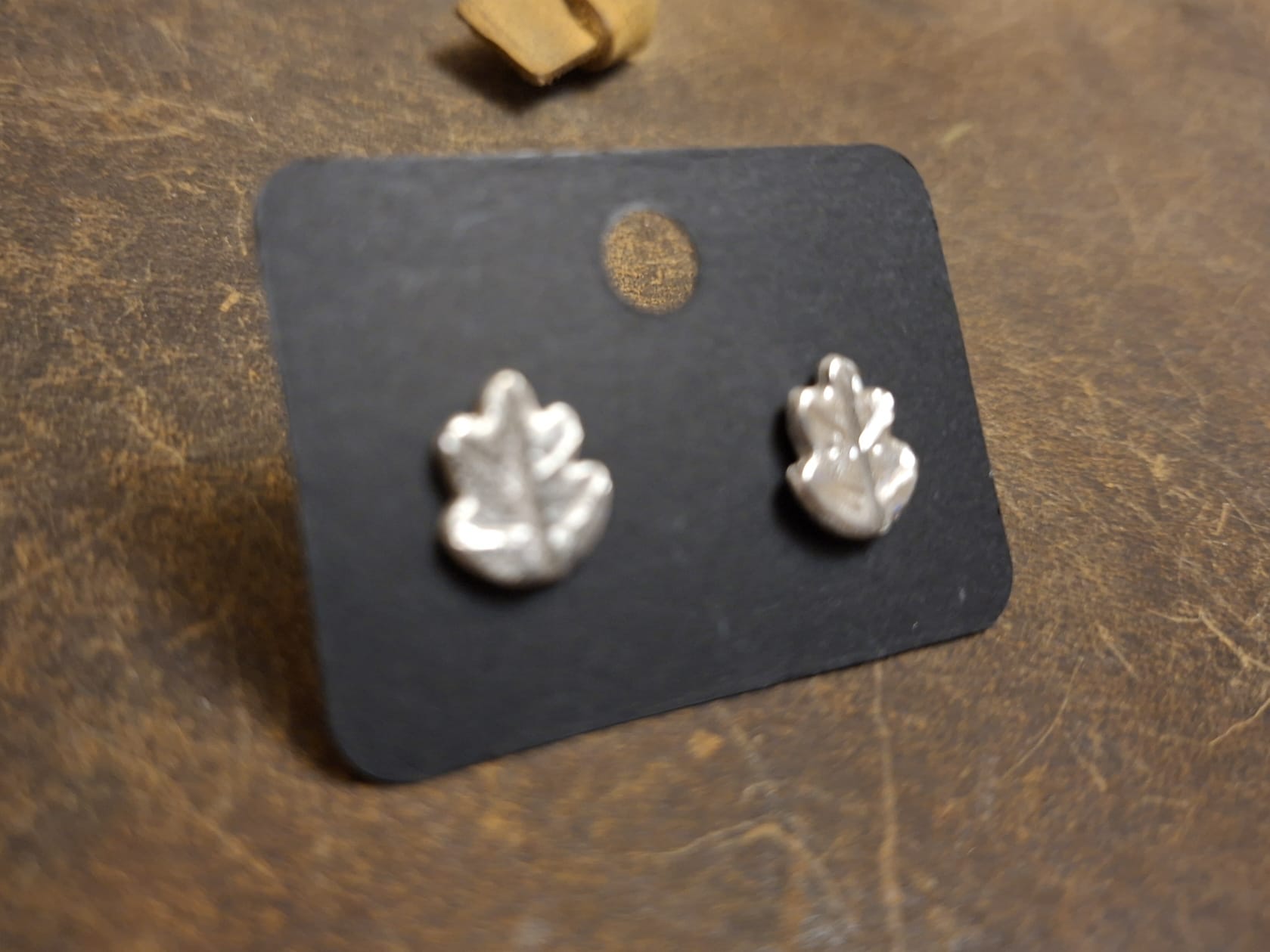 A pair of small silver leaf-shaped stud earrings displayed on a black backing card, placed on a textured brown surface.