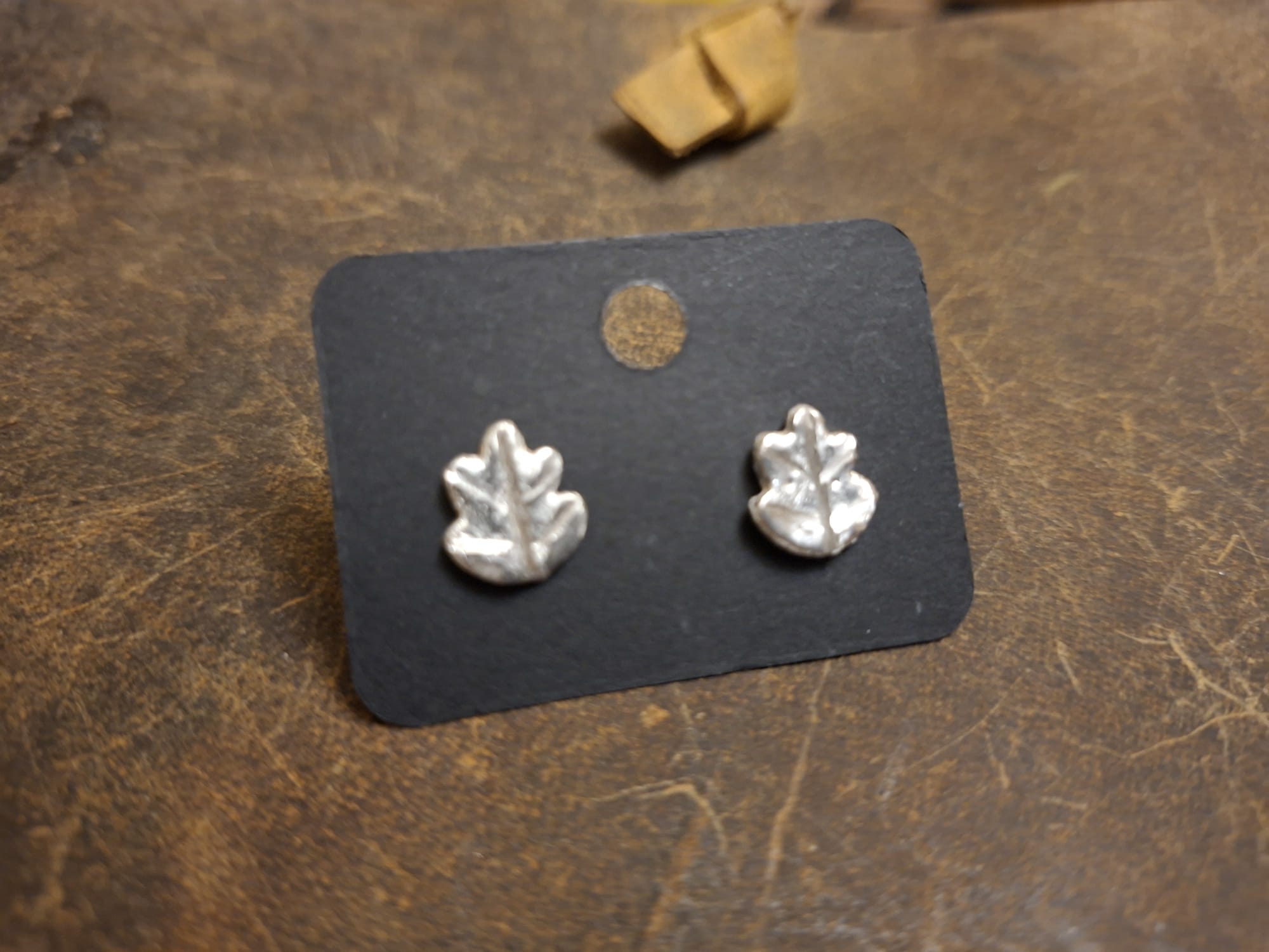 A pair of small silver leaf-shaped stud earrings displayed on a black backing card, placed on a textured brown surface.