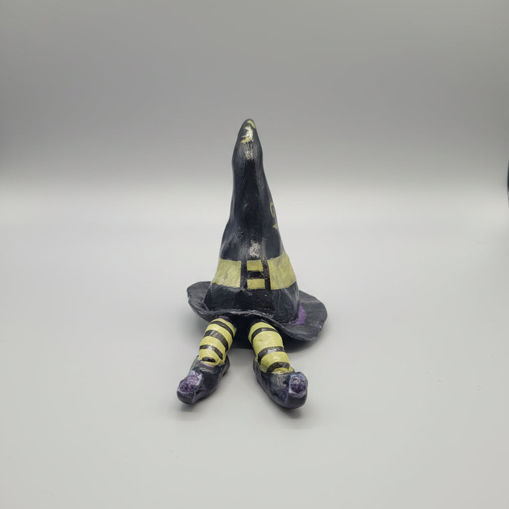 A small painted witch hat figurine with striped legs, displayed on a textured gray fabric surface in a home interior setting.