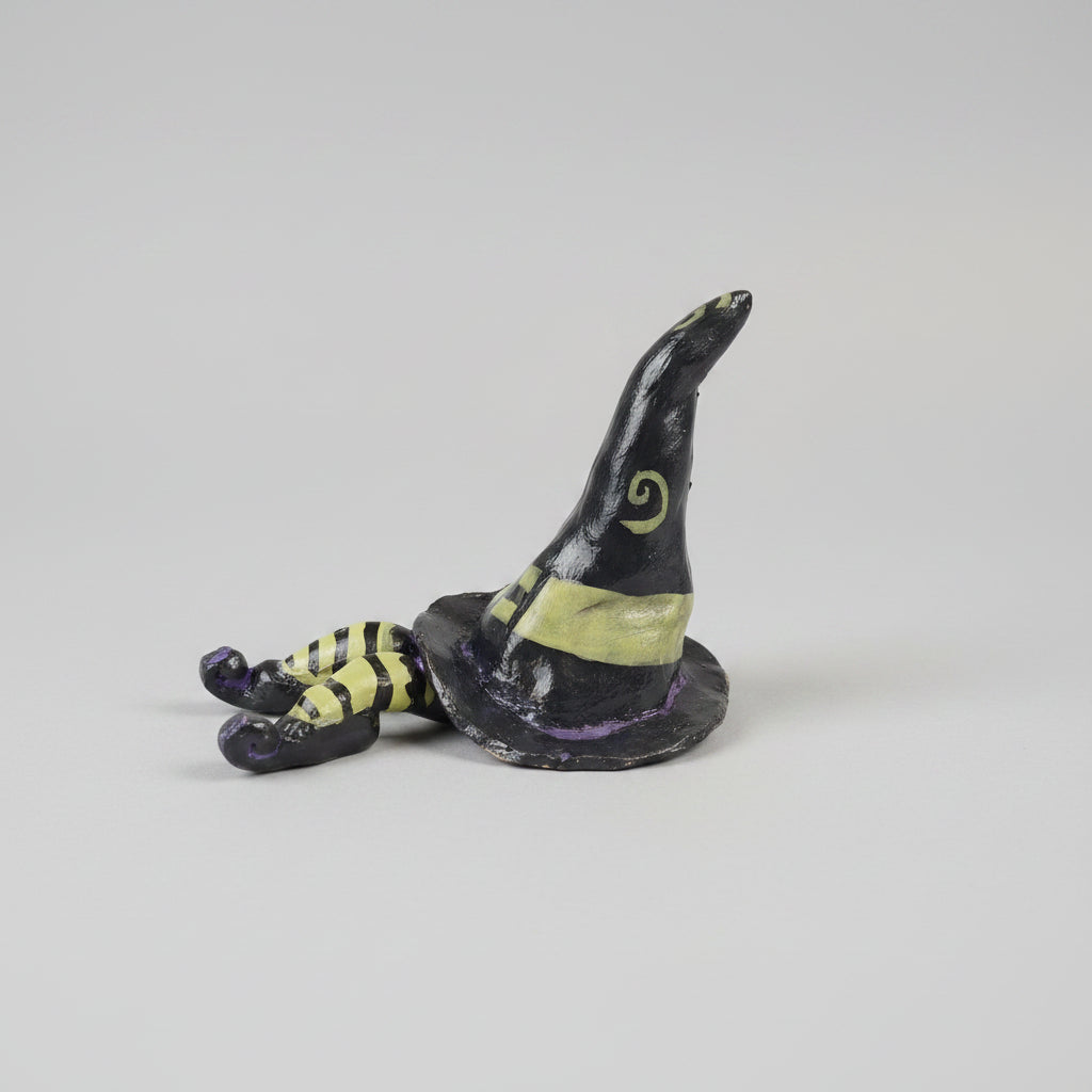 A small hand-painted witch hat figurine with striped legs, resting on a textured gray fabric surface in a home environment.