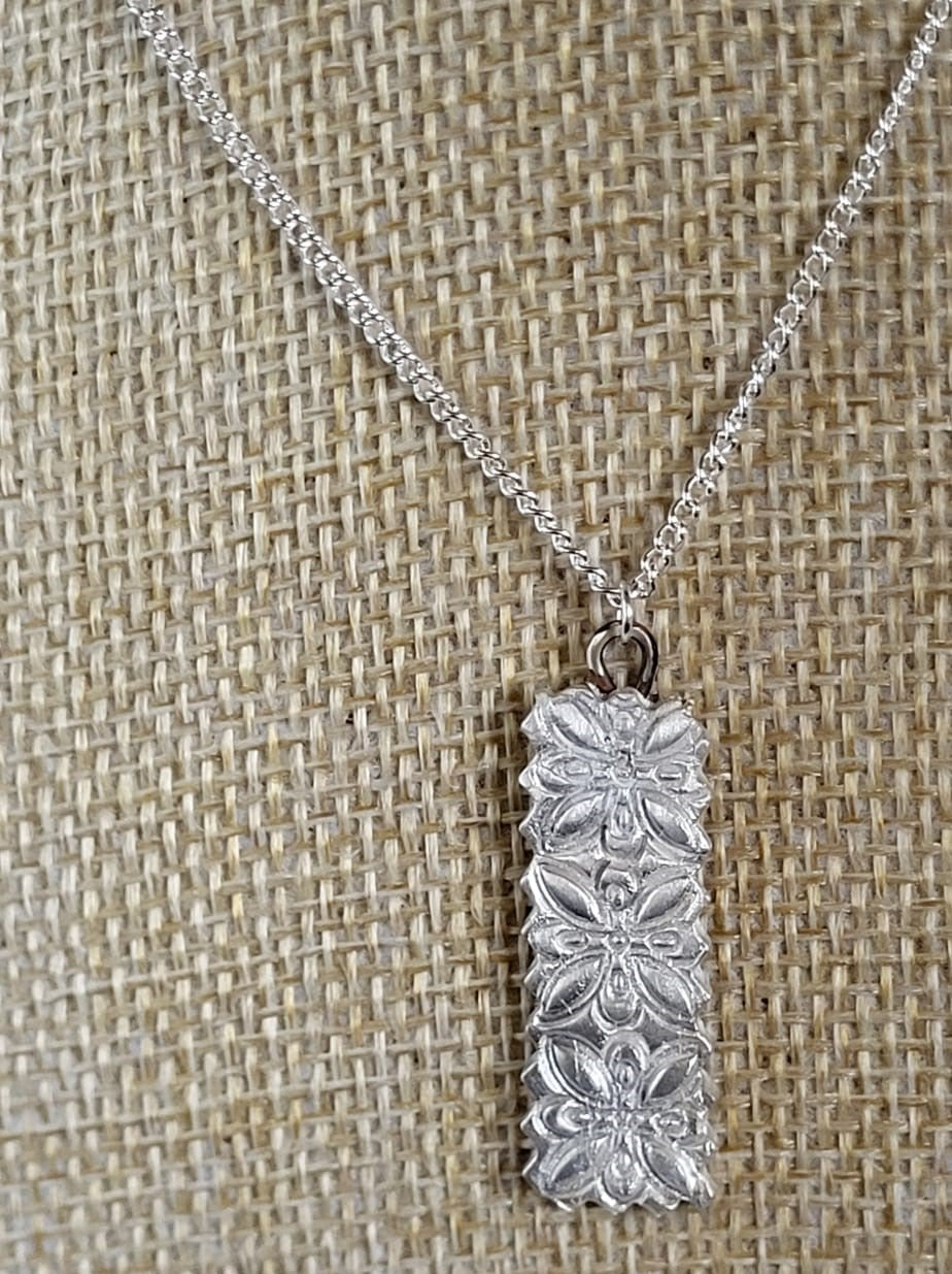 A silver rectangular pendant necklace with a floral pattern, displayed on a textured burlap background.