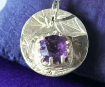 A silver pendant with a textured circular design and a central purple gemstone, displayed on a purple fabric background.