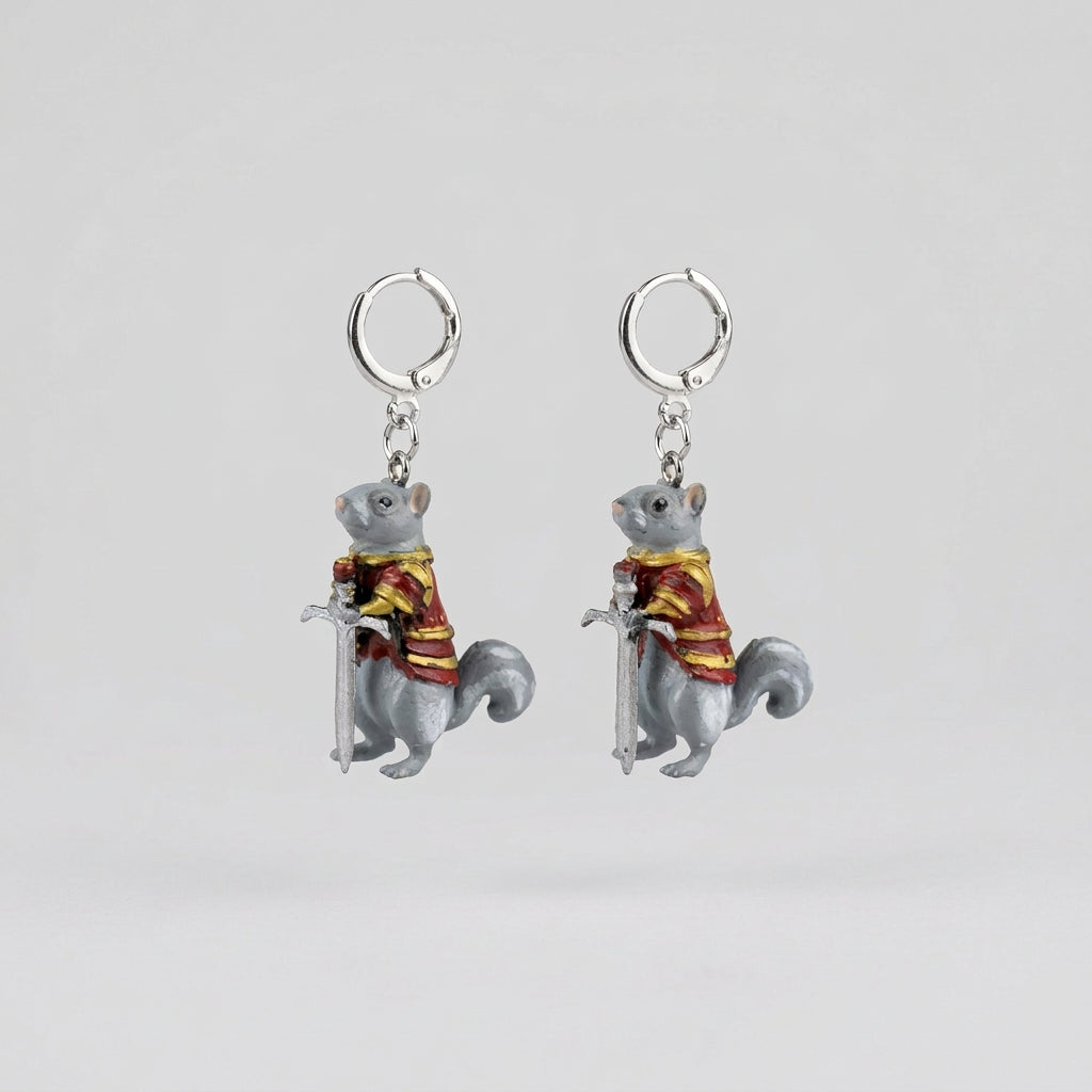 A pair of dangling earrings shaped like small gray squirrels wearing red and gold striped robes, each with a silver ring at the top, placed on a textured gray fabric surface.