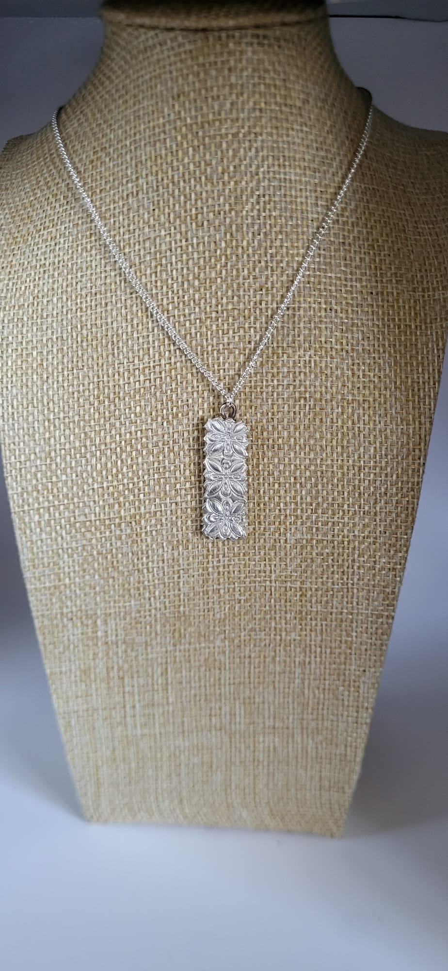 A silver rectangular pendant necklace with a textured, hammered surface, displayed on a beige burlap jewelry bust against a neutral gray background.
