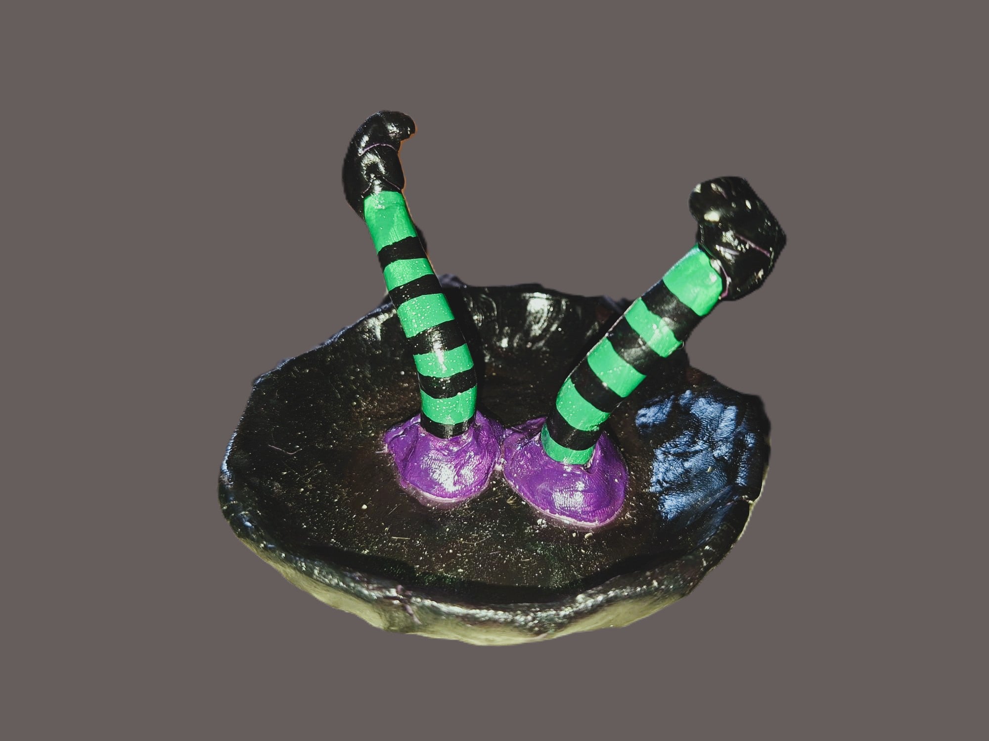 A small decorative figurine or sculpture of two striped green and black legs rising from a black base with purple feet, shown on a plain taupe-brown background.