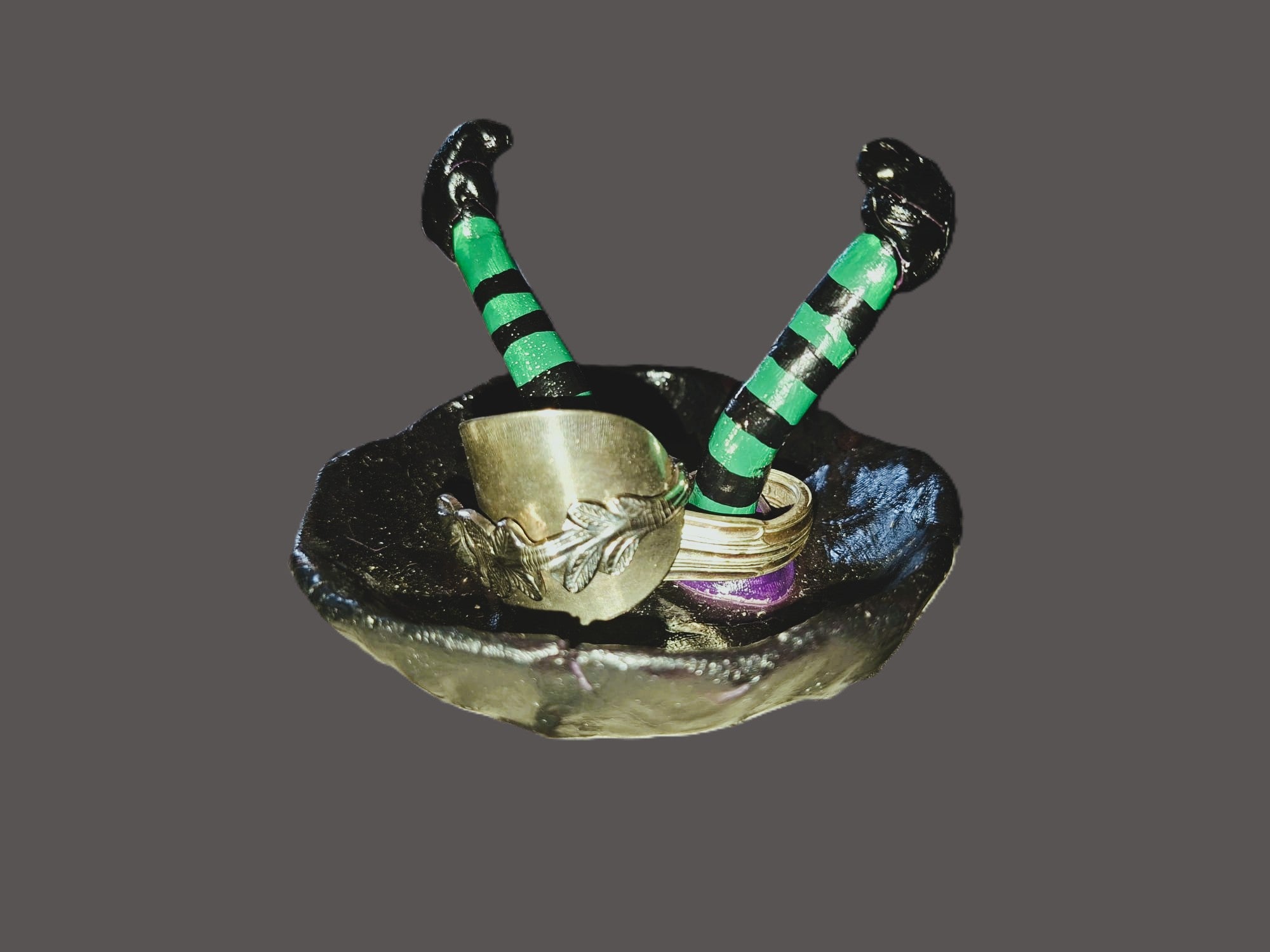 A miniature decorative figurine of a witch's scythe resting in a small black dish, featuring green and black striped handles and silver-colored blades, set against a solid dark gray background.
